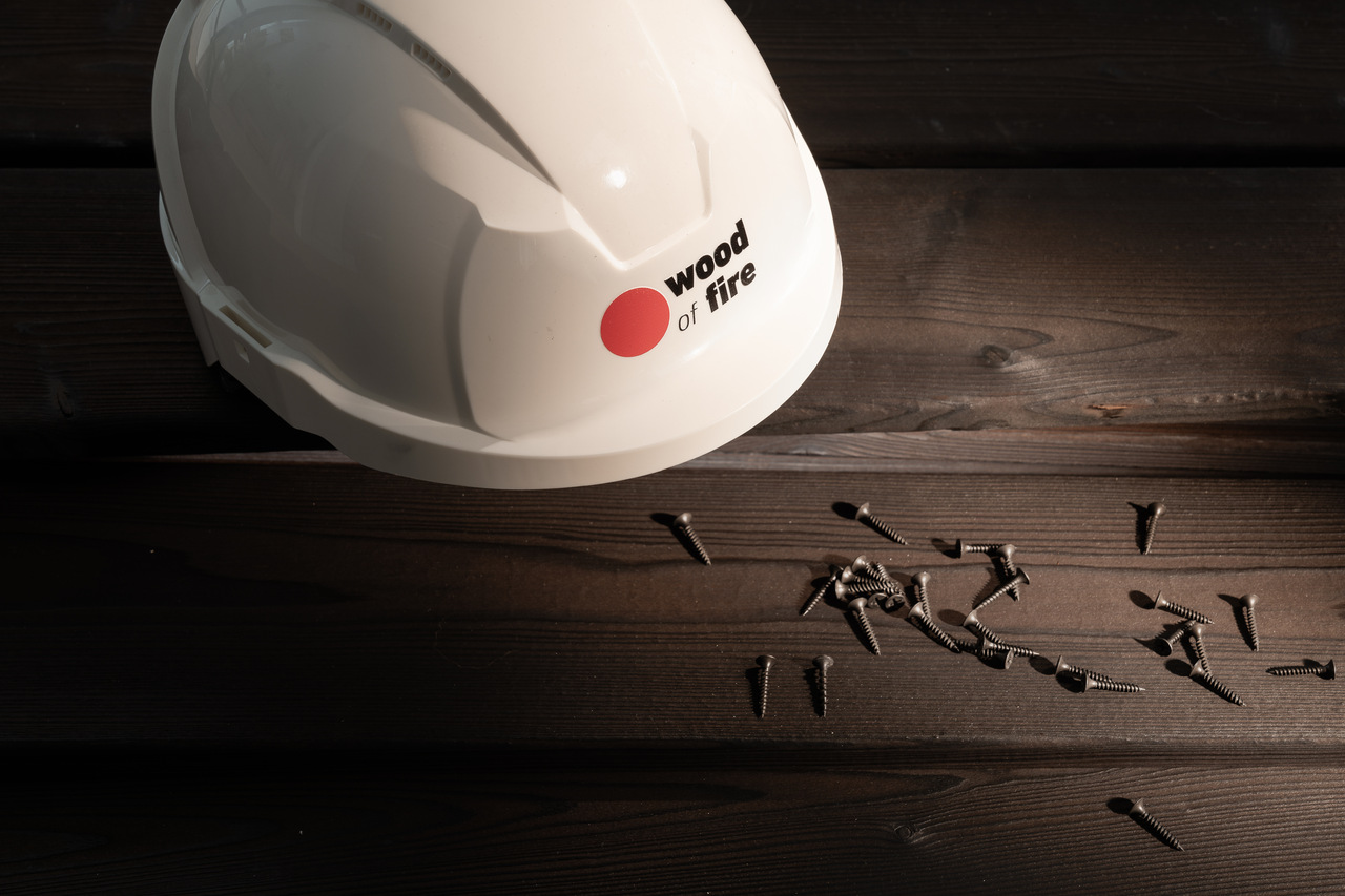 Biały kask ochronny z logo Wood of Fire leżący obok wkrętów na ciemnych deskach